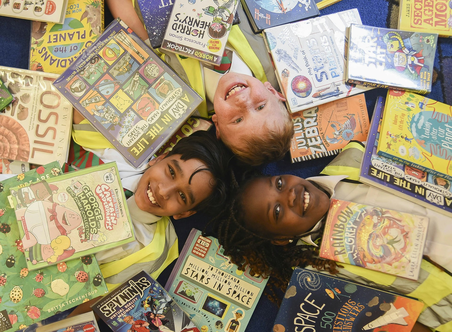 Three children lying on the floor of a library covered with books